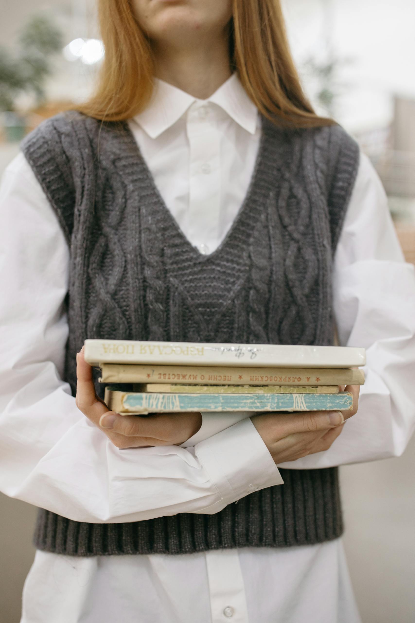 A young woman in a sweater vest holds a stack of books, emphasizing education and learning.